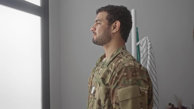 Young hispanic man in camouflage uniform with dog tags stands looking out window in building; duty reflection.