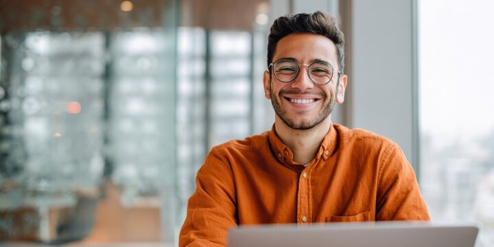 Smiling young caucasian male in orange shirt working on laptop in modern office
