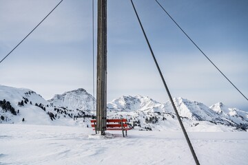 Summit cross of Simmel with rest bench in winter, overlooking snow-covered high peaks of the Bregenzerwald mountains including Braunarlspitze.