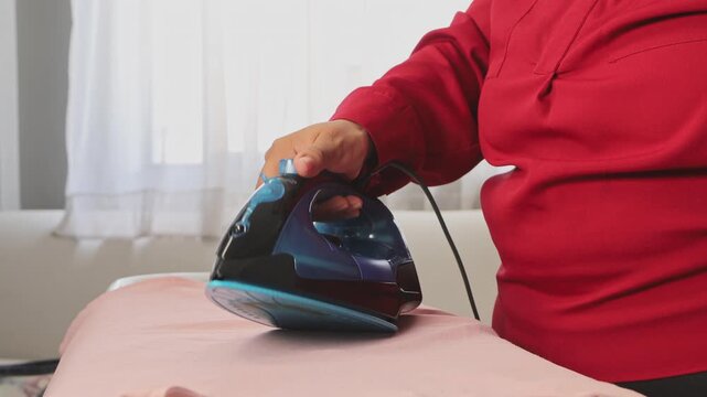 A woman carefully irons a delicate pink blouse on an ironing board, giving attention to detail to maintain the fabric&rsquo;s quality and prepare it for wearing later.