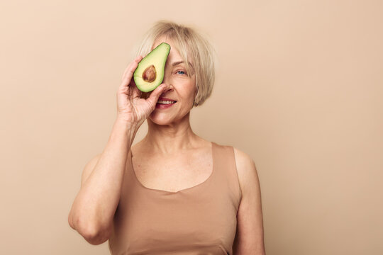Happy senior woman with short blonde hair holds half an avocado near her eye, smiling on beige background. Concept for healthy lifestyle, nutrition, and natural beauty in mature age.