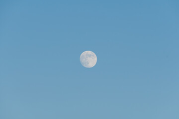 Full moon glowing in a clear, pale blue morning sky, offering a minimalist and serene celestial background for creative designs.