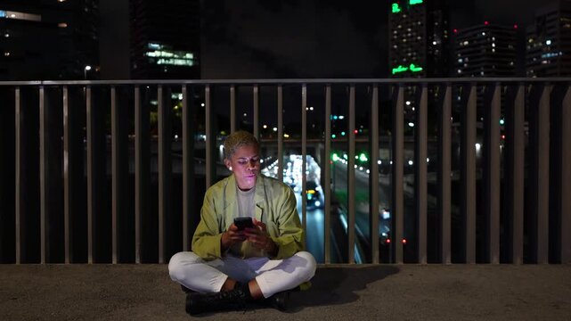Stylish non binary person sitting cross legged on a city bridge, illuminated by the smartphone screen while texting or browsing social media