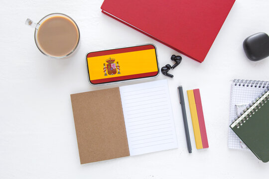  workspace with smartphone displaying Spain flag on screen, notebook, coffee cup, headphones and stationery on white desk, concept Spanish language learning, travel planning, online communication  