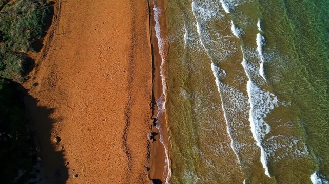 Aerial drone view of the most beautiful Malta beach in Gozo, vibrant red sand Ramla beach with crystal clear blue water and mesmerizing waves with lush green vegetation around it and the famous Mixta 