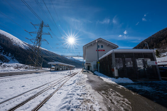 A small Zermatt alpine station sits in snowy mountains as sun flares, long shadows fall, and parallel tracks lead to a tunnel, with catenary lines and a lattice pylon.