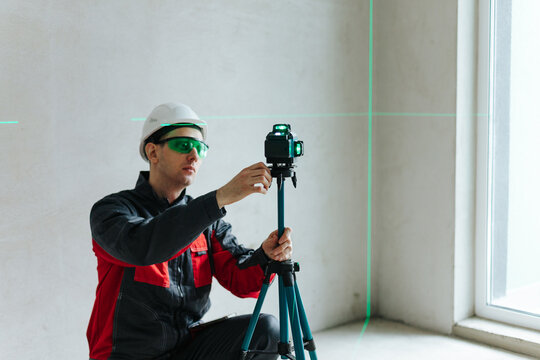 Technician Adjusting Laser Level While Wearing Safety Glasses And Helmet. Precision Alignment In Unfinished Interior Space.