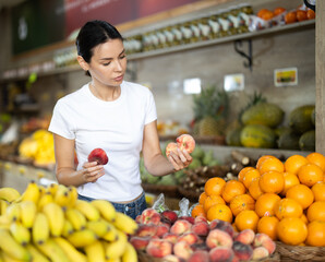 Woman stands in a supermarket near a bountiful fruit counter and chooses a flat peach. Customer prepares to weigh the fruit before buying. High quality photo © JackF