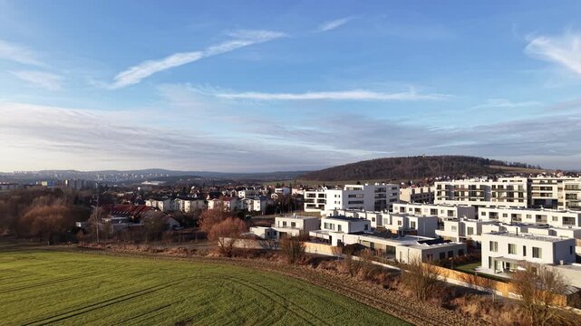 Aerial view of modern residential complex. Suburban landscape with green field and distant city skyline. Sunny day with scattered clouds. Drone footage showcasing architectural design