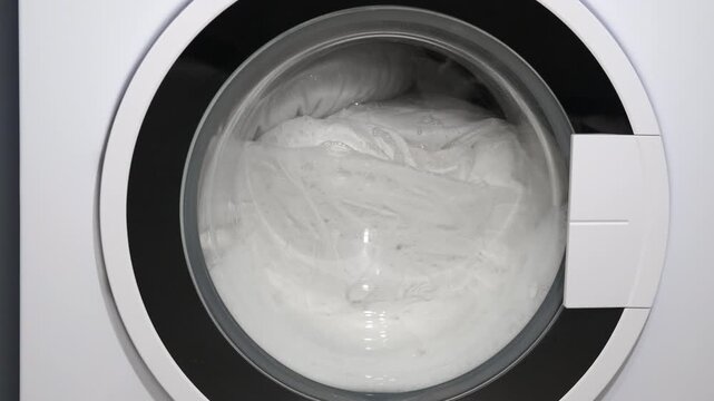 Washing machine drum spinning with white laundry. Close-up of a modern washing machine door with white clothes rotating and tumbling in soapy water during a wash cycle, representing daily housework