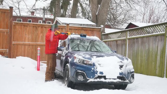 Man in winter coat brushing heavy snow off side windows of a blue SUV.