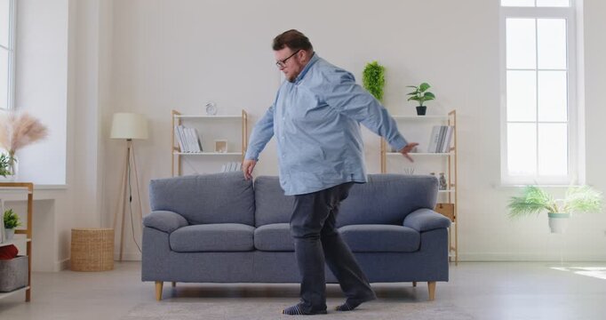 Expressive fat overweight man in glasses dancing and singing with upbeat energy near a sofa in living room, enjoying music and the moment at home, carefree. Obese male person having fun indoors.