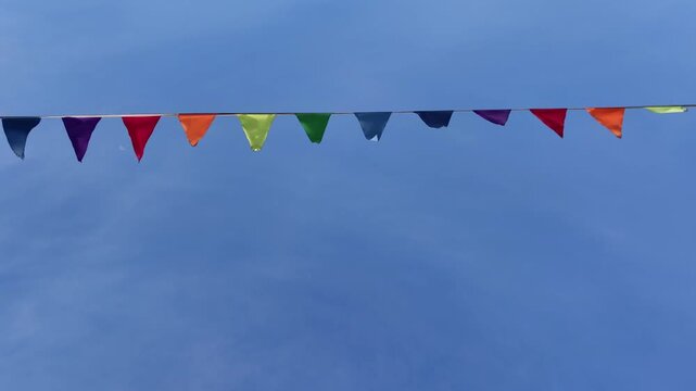colorful triangular pennant flags on string gently moving in wind against bright blue sky with light clouds, creating festive and celebratory outdoor atmosphere on sunny day. close up.