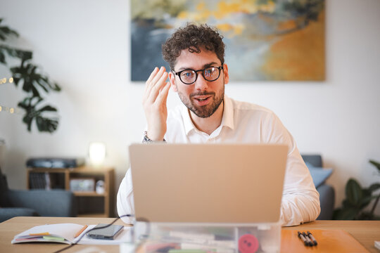 A young man with glasses and curly hair smiles while talking during a video call on his laptop, working from his home office.