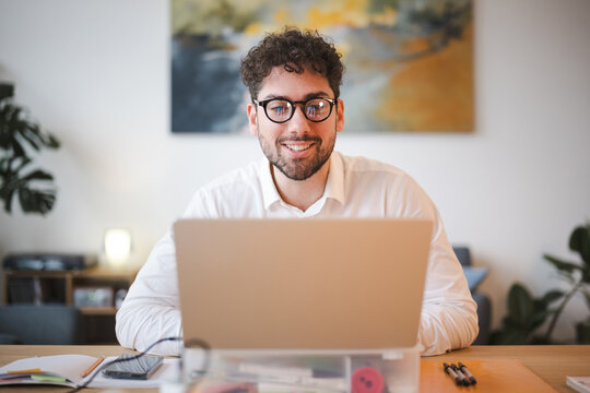 A man with curly hair and glasses smiles while working on his laptop at a desk.