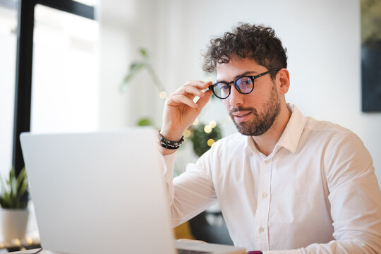 A man with curly hair adjusts his glasses while working on his laptop. He is wearing a white collared shirt and bracelets on his wrist.