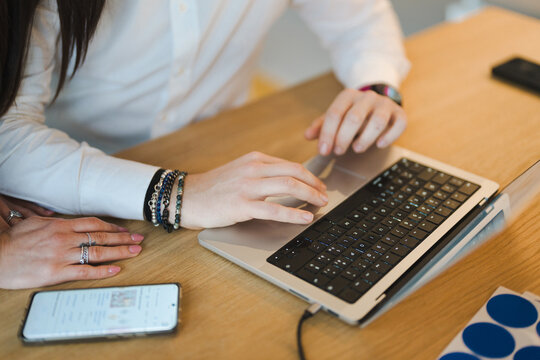 A person wearing bracelets and rings types on a laptop while a smartphone rests nearby on a wooden desk.