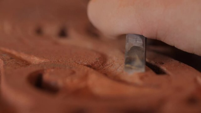 Close up macro shot of a skilled craftsman using a metal chisel tool to carefully carve and shape a rough piece of red terracotta stone in bright daylight.