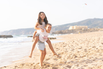 Fototapeta premium Cheerful girls playing piggyback ride on sandy beach near sea smiling and enjoying fun time during sunny day