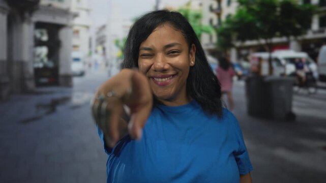 Woman wearing blue shirt smiling and pointing finger on busy city street at midday; friendly invitation.