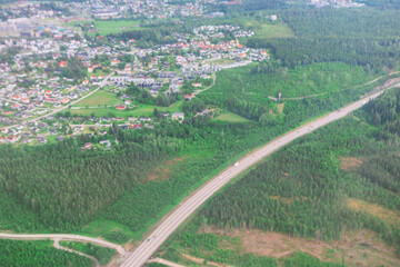 Aerial view shows a highway cutting through green forest, leading toward a suburban area with houses and open fields. Landscape blends natural greenery with developed neighborhoods