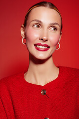 Smiling woman with red lipstick and hoop earrings wears red sweater against red background. Close-up portrait with natural skin glow and confident expression. Concept of beauty and fashion. © SHOTPRIME STUDIO