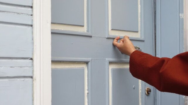 Woman's hand knocking a vintage door knocker on a gray wooden door with white trim, close-up shot capturing dynamic motion and detail