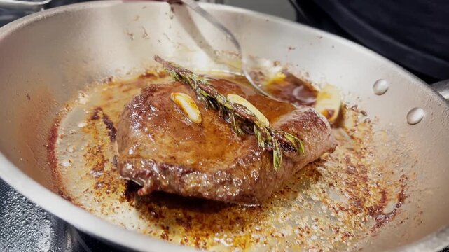 Juicy beef steak frying in stainless steel pan with garlic and rosemary. Close up of chef basting steak with hot butter using spoon. Delicious gourmet cooking process in kitchen.