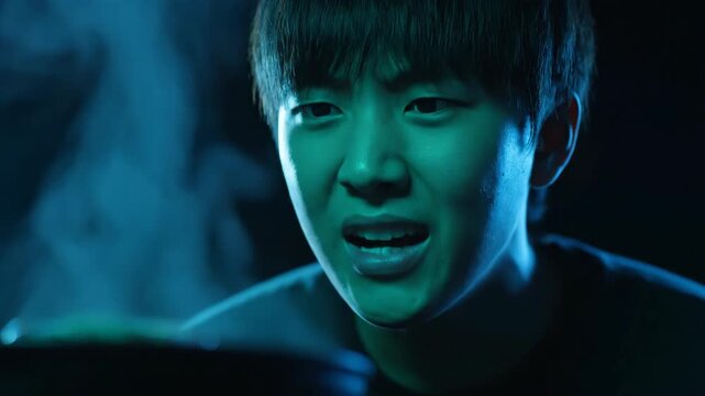 Young man looks surprised at steaming cauldron in dark, with blue and green lighting and smoke, magic concept.