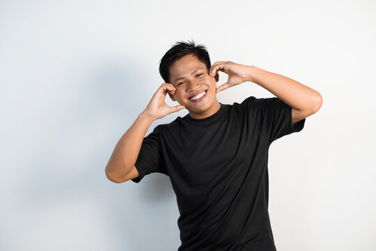A cheerful young Asian man wearing a black shirt expresses happiness with a heart shaped hand gesture placed on his cheek against a white background.
