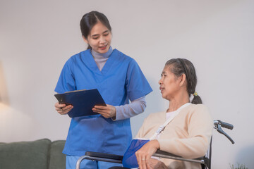 Obraz premium A young Asian nurse discusses medical information with an elderly female patient seated in a wheelchair with an arm sling at home, highlighting professional healthcare support, rehabilitation