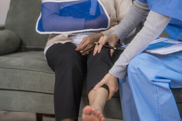 Obraz premium A young Asian nurse discusses medical information with an elderly female patient seated in a wheelchair with an arm sling at home, highlighting professional healthcare support, rehabilitation