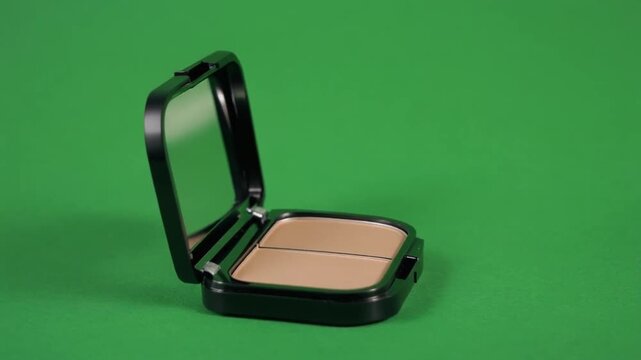 Open black compact makeup case with two-toned beige powder on a green background, product photography with studio lighting.