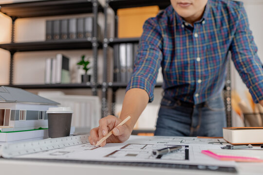 A young architect works with building plans and a house model in the office, reviewing blueprints, marking details, and analyzing design structures with focus and creativity.
