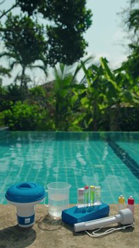 Swimming pool maintenance equipment with chlorine dispenser, water test kit, measuring cup and chemical reagents on pool edge, private pool water treatment concept with tropical garden background 