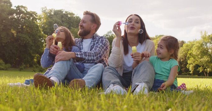 Outdoor view of happy mother, father, daughter and son blowing soap bubbles while sitting on green grass in park, enjoying summer holidays and spare time. Parents playing with children 