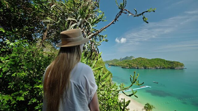 Young woman in a hat standing on a tropical viewpoint, looking at emerald lagoon and limestone islands in Ang Thong Marine Park, Thailand, surrounded by lush green vegetation and cactus plants