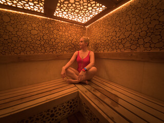 Young woman in pink swimsuit sitting in luxury sauna with decorative wooden log walls and ornate glowing geometric ceiling patterns