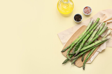 Wooden board with fresh green asparagus and jug of oil on yellow background