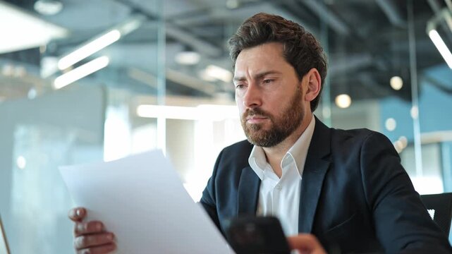 Professional compares crucial business documents with information on smartphone. Serious demeanor and focused attention indicate deep thought and analytical review of important data in office.