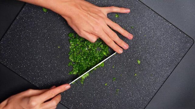 Person mincing fresh green cilantro on dark board. Top view of hands using a knife to finely chop green coriander herbs on a black cutting board.
