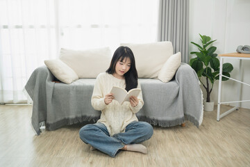 Obraz premium Woman reading a book while sitting on the floor in a cozy living room with a comfortable couch and houseplants, enjoying a moment of relaxation and inner peace