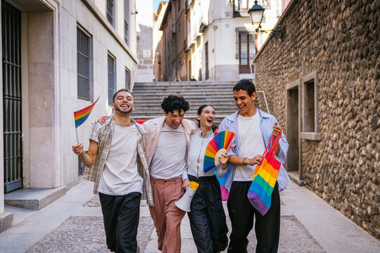 Four happy diverse friends walking together in a city street, carrying rainbow flags and a fan, celebrating lgbtq plus pride