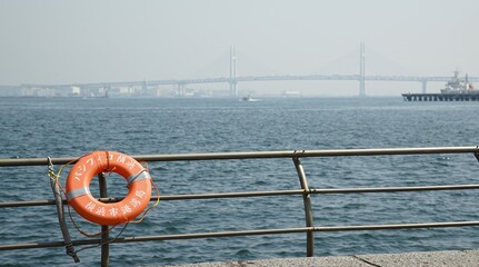 Naklejka premium An Orange Lifebuoy Hanging on a Metal Railing Overlooking the Sea with a Large Bridge in the Distance