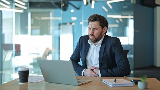 Professional experiences sudden, severe abdominal pain while working at laptop in modern office. Discomfort distress are evident. Feelings of illness and suffering interrupt focus, need for relief.