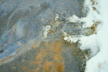 Oil slick displaying vibrant rainbow colors on dark asphalt, mixing with dirty melting snow and boot prints, showcasing concrete pollution