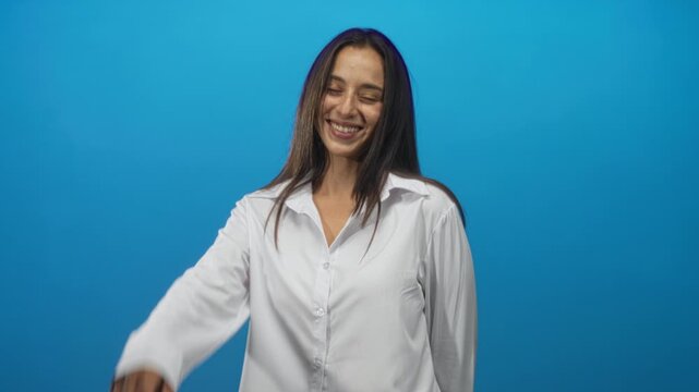 Woman smiling with eyes closed and hand reaching forward wearing white shirt and long dark hair in blue studio; joy.