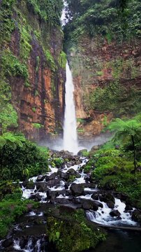 Vertical drone view of Kapas Biru Waterfall in East Java showing a tall powerful stream falling into a rocky river, surrounded by dense tropical jungle and misty canyon walls.