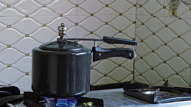 On the gas stove, a pressurized black cooker releases a steady, high-pitched hissing sound. Plumes of white steam puff from the metal whistle, filling the kitchen with the noisy rhythm of home cooking