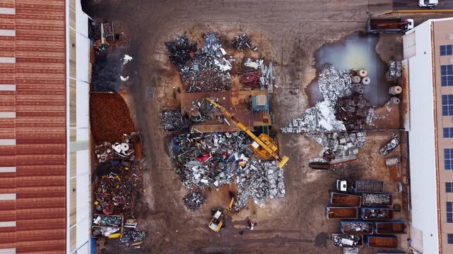 Top down aerial of an industrial metal recycling yard showing sorted scrap piles, excavator handling material, containers, drainage areas, and warehouse rooftops